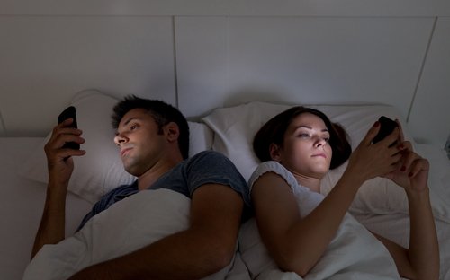 Par der ligger i sengen med hver deres telefon