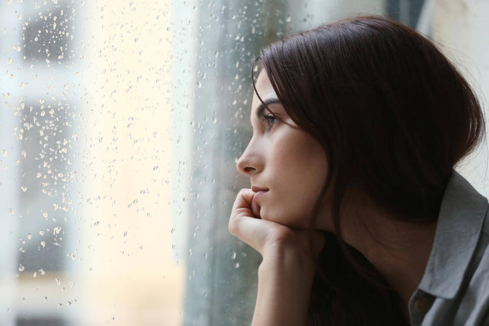 Kvinde der stirrer ud af et vindue med regn paa