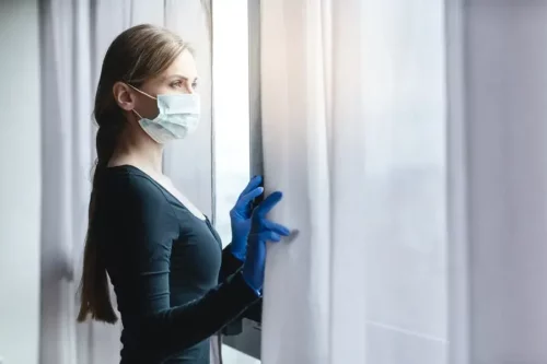 Kvinde med mundbind og handsker på ser ud af vindue
